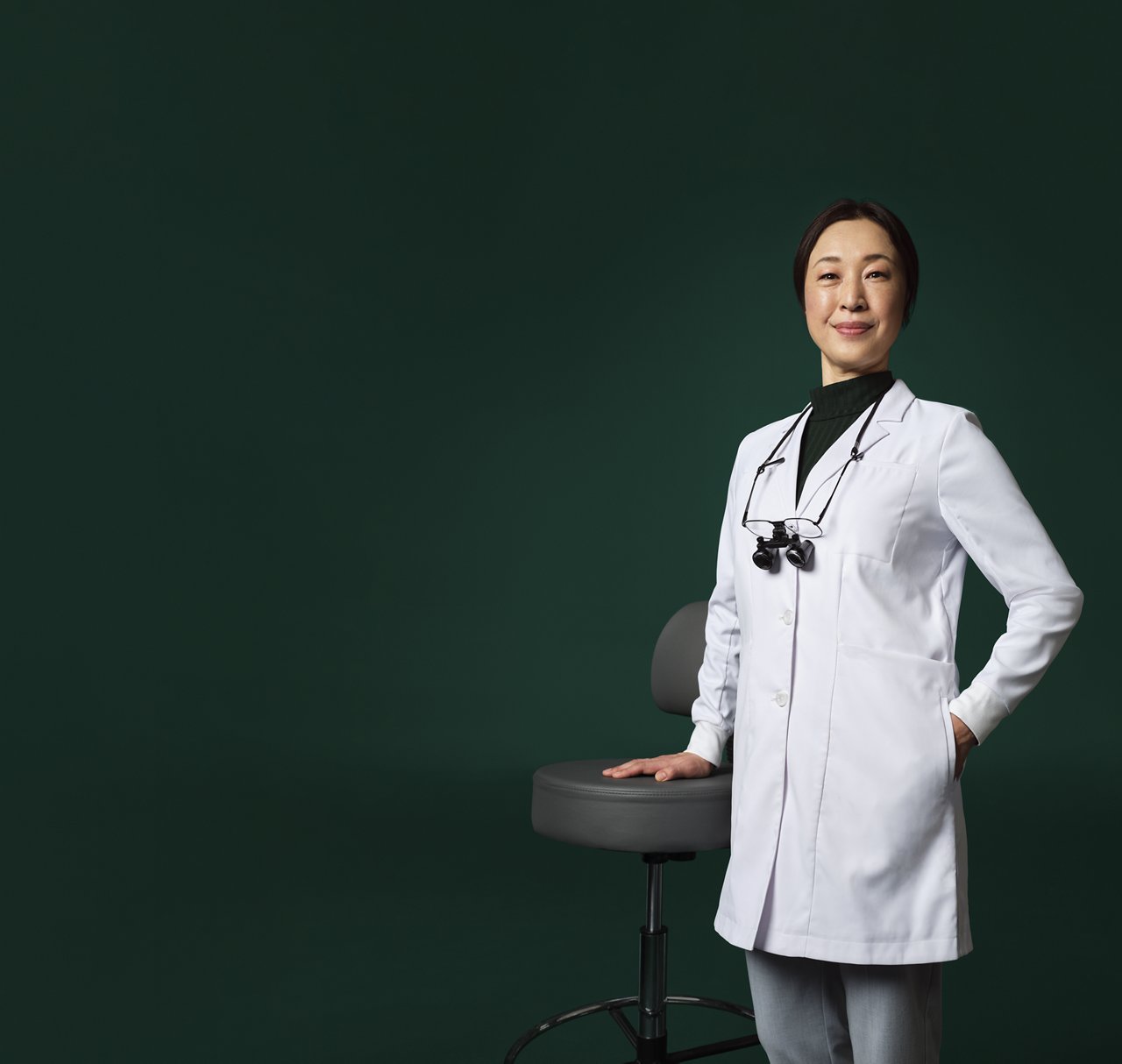 Dentist wearing loupes around their neck, in a white coat, standing with one hand on a stool.