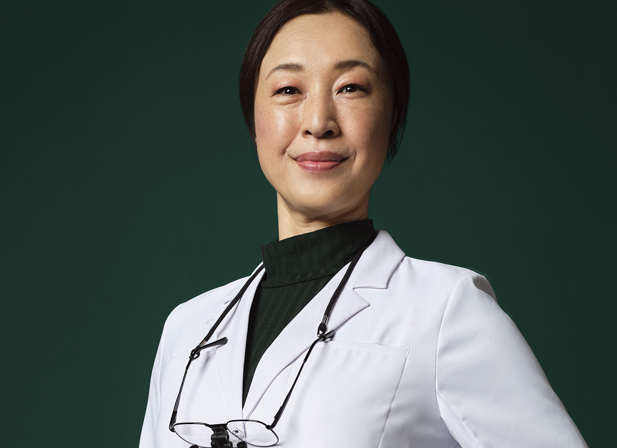 Dentiste avec des loupes autour du cou, en blouse blanche, se tenant avec la main sur un tabouret.