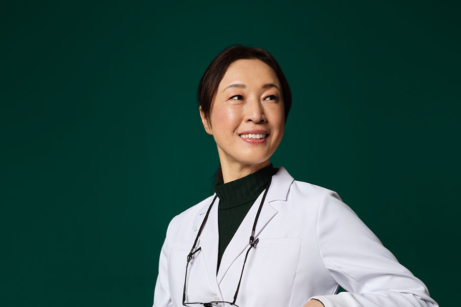 Une femme en blouse de laboratoire avec des lunettes et une loupe attachée autour du cou s’assoit et sourit.