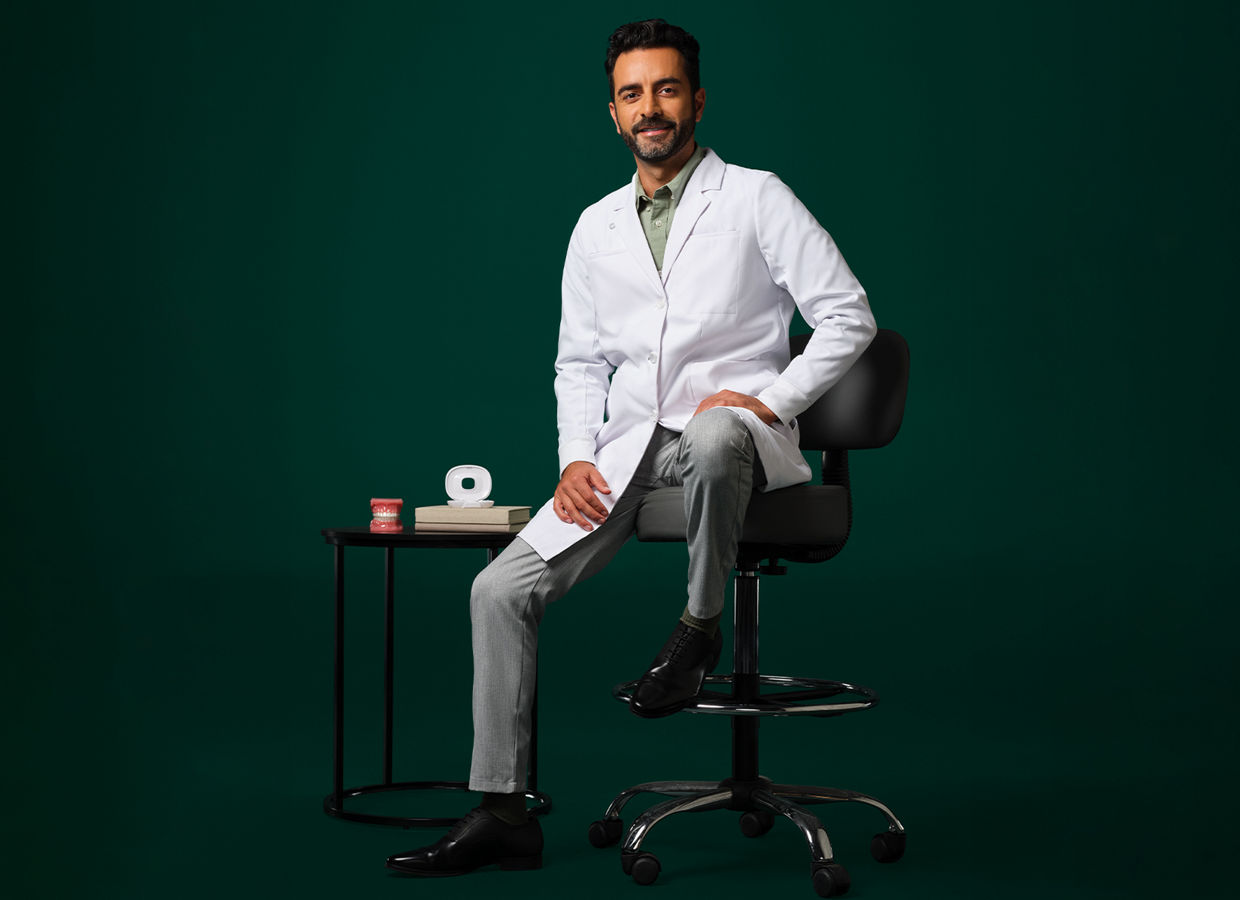 Orthodontist in a white coat sitting on a stool next to a small table displaying an aligner case and typodent.