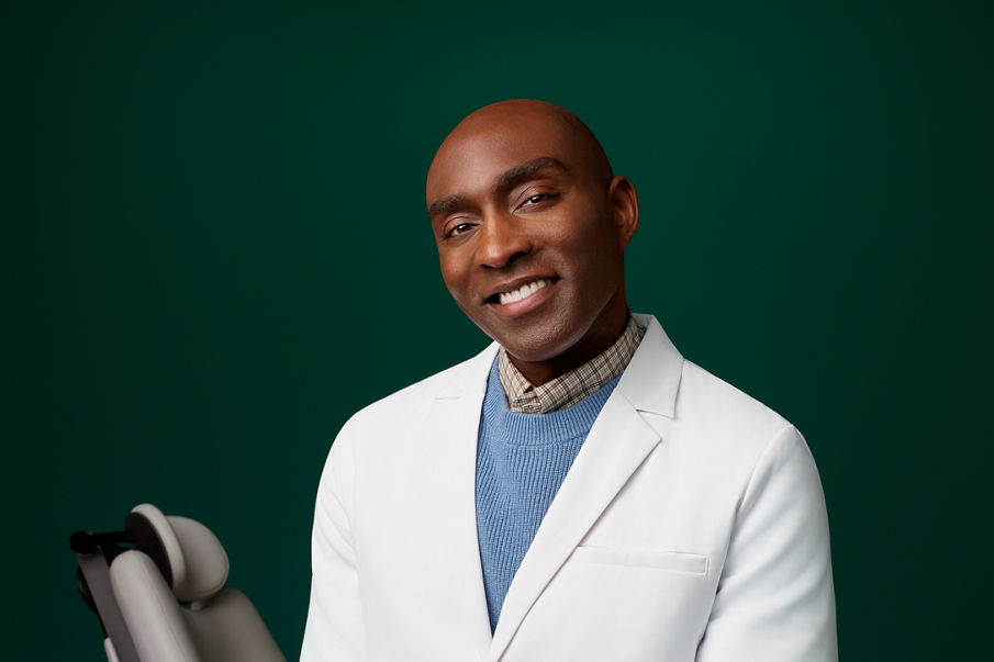 Portret van een mannelijke orthodontist, zittend naast de tandartsstoel van een patiënt, tegen een donkergroene achtergrond.