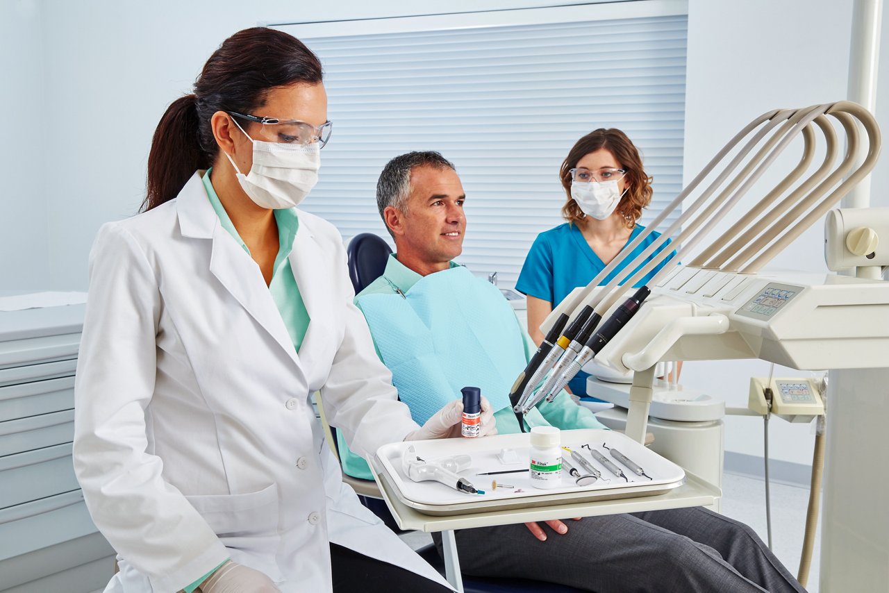 Doctor, paciente y asistente dental en la sala de consulta, doctor sosteniendo una botella de Adhesivo Universal Plus 3M™ Scotchbond™, Teal de Solventum aplicado a las camisas y la botella del producto SBU Plus reemplazada en la mano. Imagen de personas, Aplicación del producto, RGB, TIF