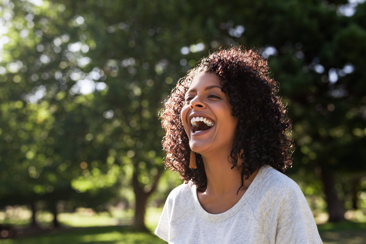 Young woman with curly hair laughing while standing outside in a park on a sunny summer afternoon; Shutterstock ID 1687578475; purchase_order: credit card; job: Brand Design - Mark Bonesteel; client: mwbonesteel1@mmm.com; other: USA