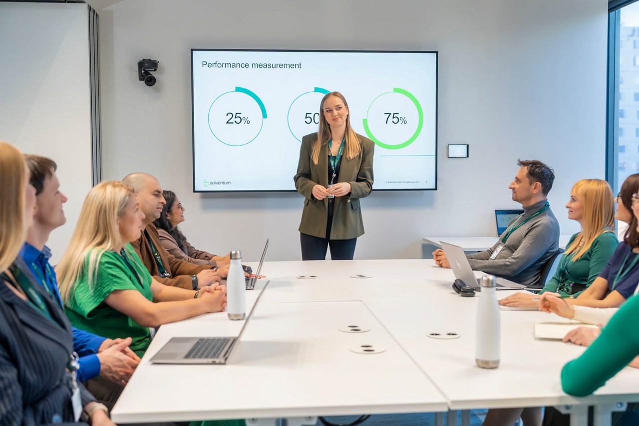 A team of Solventum employees sit around a conference table while a presenter stands and displays performance metrics on a large screen in a modern meeting room.
