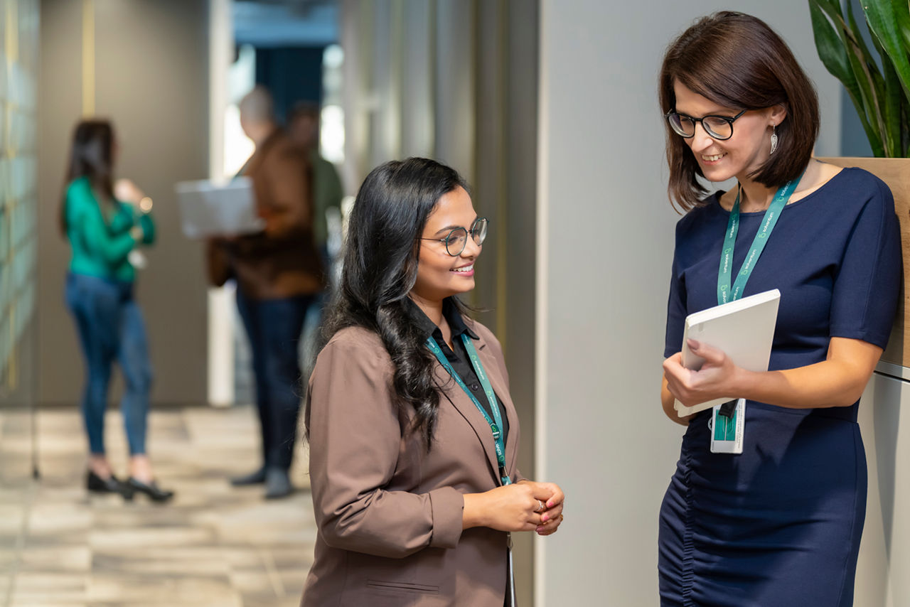 Two female Solventum employees wearing lanyards converse in a modern office hallway while holding documents and tablets, with other colleagues in the background.